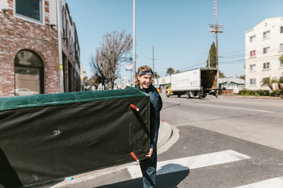 A person with shoulder-length hair wearing a black headband, black jacket, and jeans is standing on a city street corner, carrying a large, rectangular piece of outdoor furniture or equipment with a black fabric covering and green cushioning on top. The person is smiling and appears to be in the process of moving or discarding the item, which could be part of a rubbish clearance or private disposal task. In the background, a delivery truck with an open cargo area is visible driving along the road, while multi-storey brick and white residential buildings line the street. The scene is illuminated by daylight, with shadows cast on the pavement and surrounding structures, indicative of clear weather. The urban environment includes a traffic light pole, utility wires, and a few leafless trees, providing context for an outdoor rubbish removal or clearance activity managed by professional waste management services such as Garbage Disposal, which may facilitate alternative waste handling or on-site clearance tasks in urban settings.