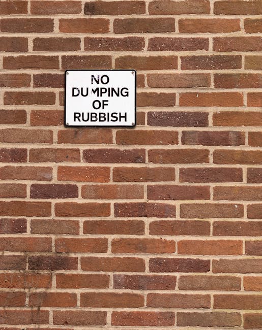 A close-up view of a brick wall featuring a rectangular metal sign with a white background and black text that reads 'NO DUMPING OF RUBBISH', mounted using four screws at each corner. The wall is constructed of uniformly arranged reddish-brown bricks with darker and lighter variations, mortar joints visible between bricks, and a slightly textured surface. The lighting is natural, casting minimal shadows, and the sign is positioned centrally within the frame, indicating an area where private rubbish removal or on-site clearance may be restricted to prevent illegal dumping, reflecting the importance of proper disposal practices often managed by waste removal service providers like Garbage Disposal.