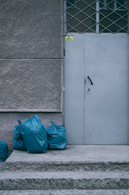 The image shows three blue rubbish bags made of plastic, placed on a concrete step in front of a grey door and a textured grey wall. The bags appear full and sealed, with some gathered at the top. The door is metallic, painted in a light grey color, and features a black handle and lock. Above the door, there is a section of metal grille or louvered vent, indicating ventilation or security measures. The surrounding environment is an exterior urban setting, with the bags positioned against the building's façade, possibly awaiting collection or removal. The scene is well-lit with natural daylight, highlighting the textures of the concrete, the plastic bags, and the building surfaces. This visual context aligns with services such as private rubbish collection or alternative waste handling, often managed by companies like Garbage Disposal, emphasizing the importance of proper waste management and disposal outside standard municipal collection points.
